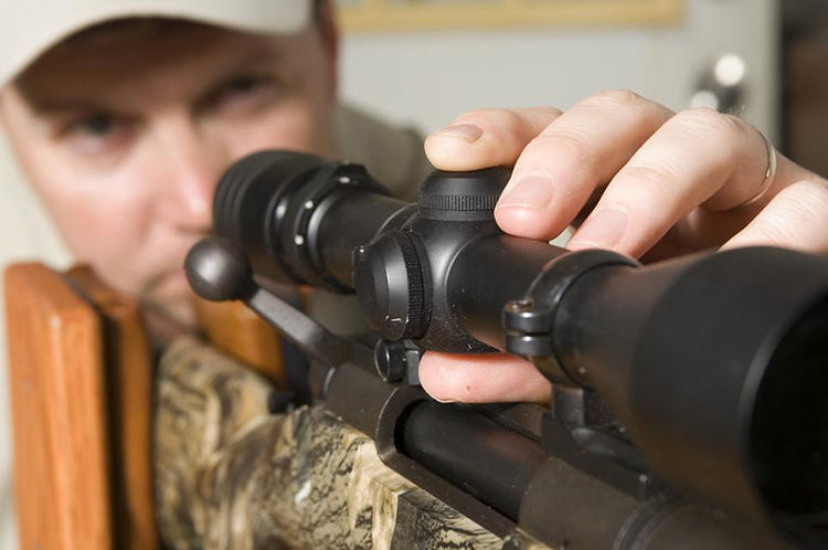 Person adjusting the scope on a camouflaged rifle while looking through it.
