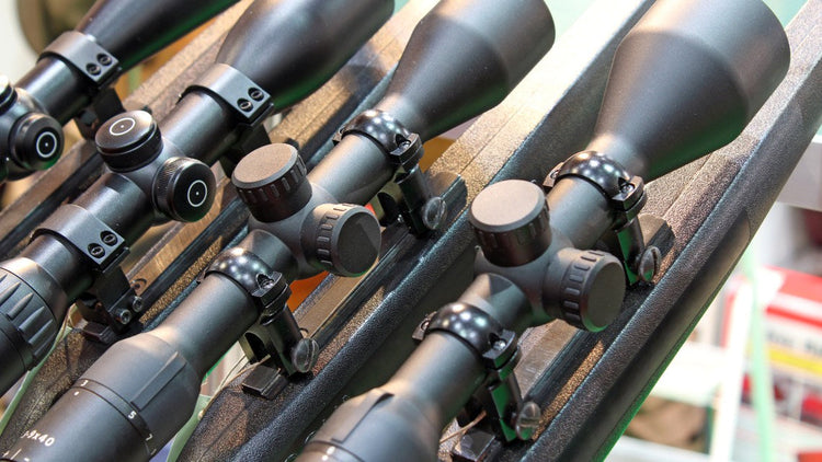 Multiple rifle scopes displayed in a row, mounted on a rack, showing adjustment knobs and lenses.